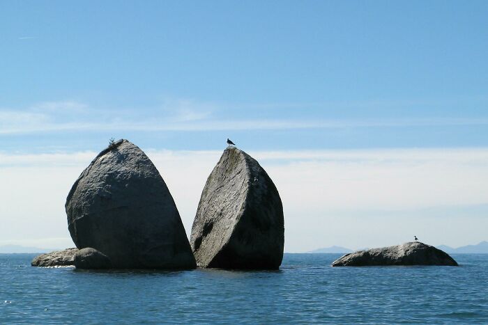 Split Apple Rock, a large boulder cut in half, sits in clear blue water with a bird perched on top, a surprising photo of an object cut in half.