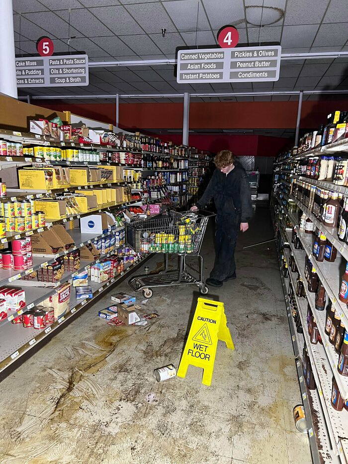 A person exploring abandoned places, standing in a disheveled supermarket aisle with a shopping cart, wet floor sign, and scattered items, finding something very unexpected.