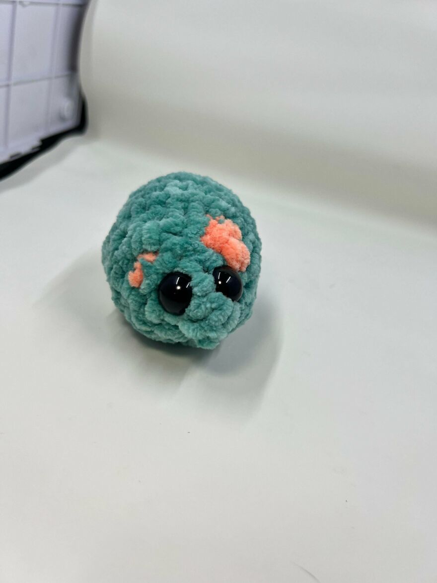 A small, teal and orange crochet creation with large black eyes on a white background, proving not every craft project turns out as intended.