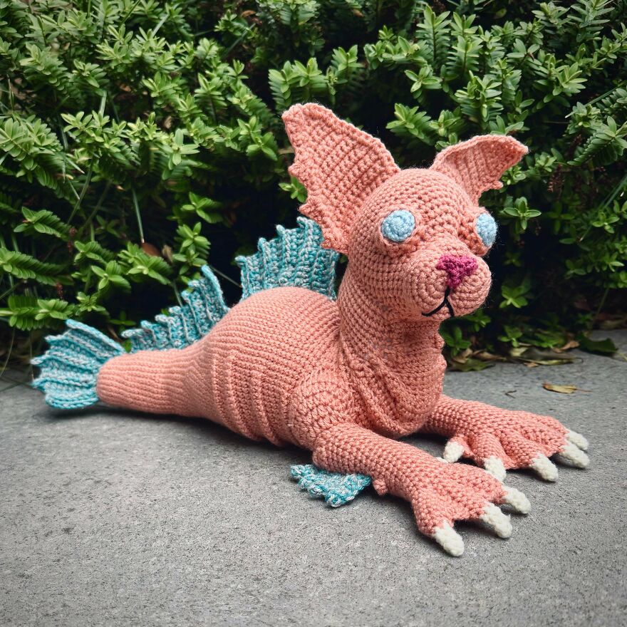 A pink crochet creation with blue fins and white claws, resembling a mythical creature, outdoors in front of green foliage.