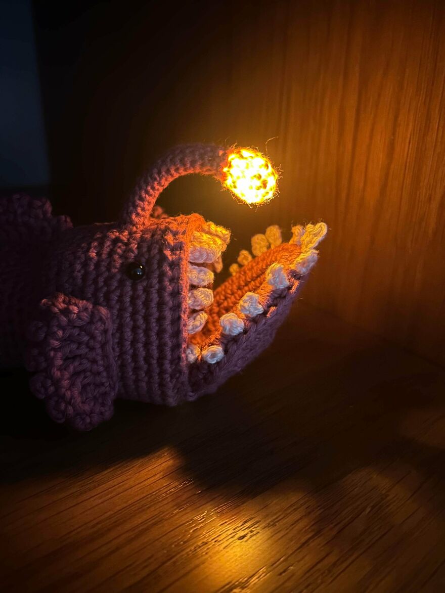 A purple crochet creation of an anglerfish with glowing lure and sharp teeth in a dimly lit setting.