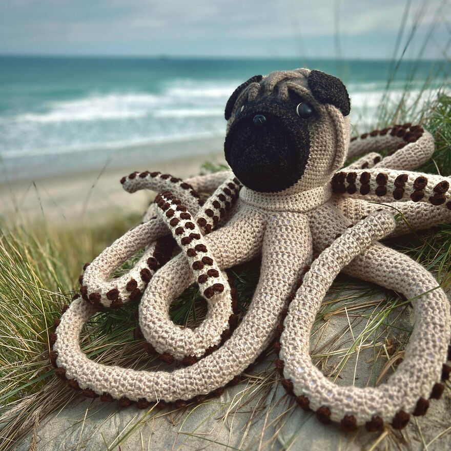 A large crochet creation, a pug-octopus hybrid, sitting in the sand with tall grass, overlooking the ocean.