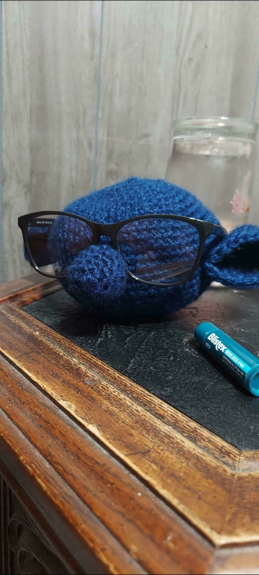 A blue crochet creation wearing eyeglasses, a humorous craft project on a wooden table.