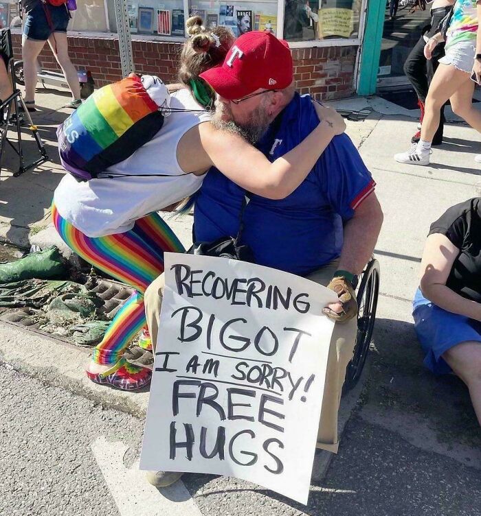 A person hugging an elderly man in a wheelchair holding a sign: Recovering Bigot, I am sorry! Free Hugs, showing character development.