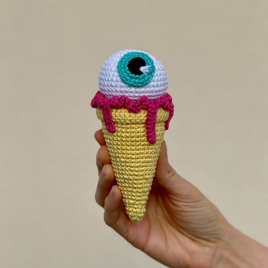 A hand holds a spooky crochet ice cream cone with an eyeball scoop and pink syrup, a funny craft project.