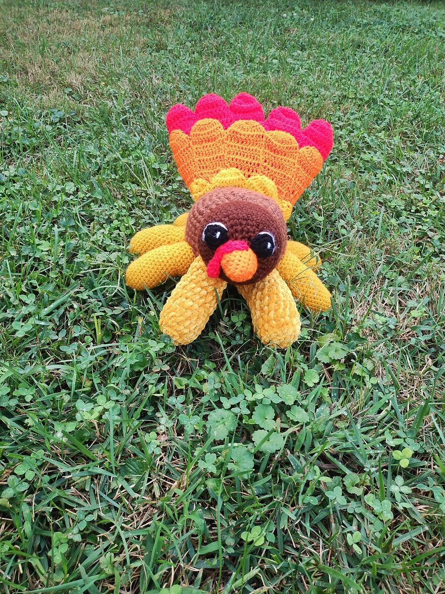 A funny crochet turkey with yellow body, orange and red tail, and brown head, laying in green grass.