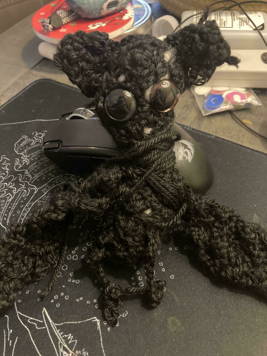A small, black, multi-limbed crochet creation with large dark eyes sits on a desk. Crochet craft projects.