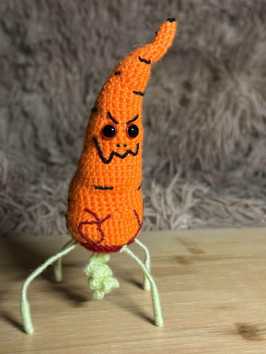 An orange crochet creation, a carrot with an angry face and root-like legs, standing on a wooden surface.