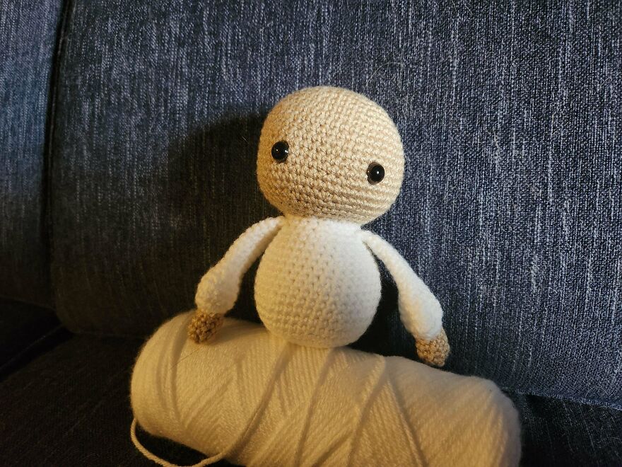 A small, tan and white crochet creation doll, resembling a human figure, sitting on a skein of yarn.