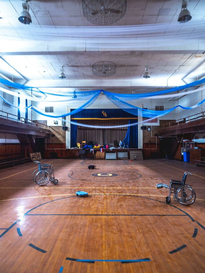An abandoned school gym with a stage, basketball court, and two wheelchairs, decorated with blue and white fabric, found while exploring abandoned places.