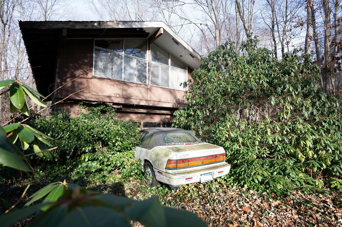 An abandoned car overgrown with foliage sits in front of a modern-style, decaying abandoned house, found while exploring abandoned places.