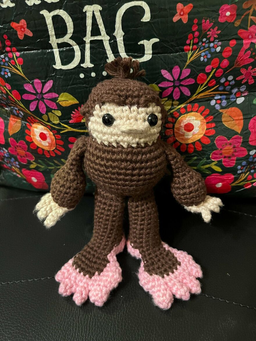 A brown and cream crochet creation doll with pink feet, standing in front of a colorful bag.