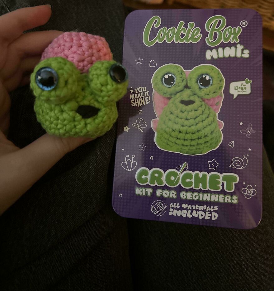 A small green and pink crochet creation frog from a beginner's kit next to its packaging.