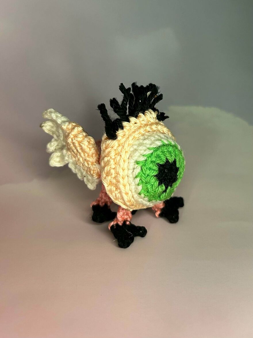 A small, whimsical crochet creature with a large green eyeball, black hair, and tiny wings, showcasing a unique craft project.