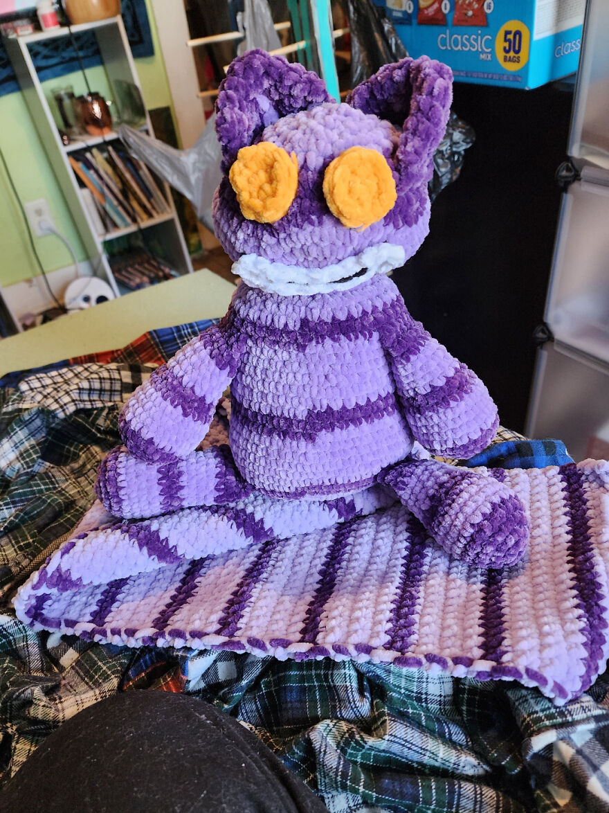 A plush purple and light purple striped crochet cat with large yellow eyes sits on a plaid blanket, a charming crochet creation.