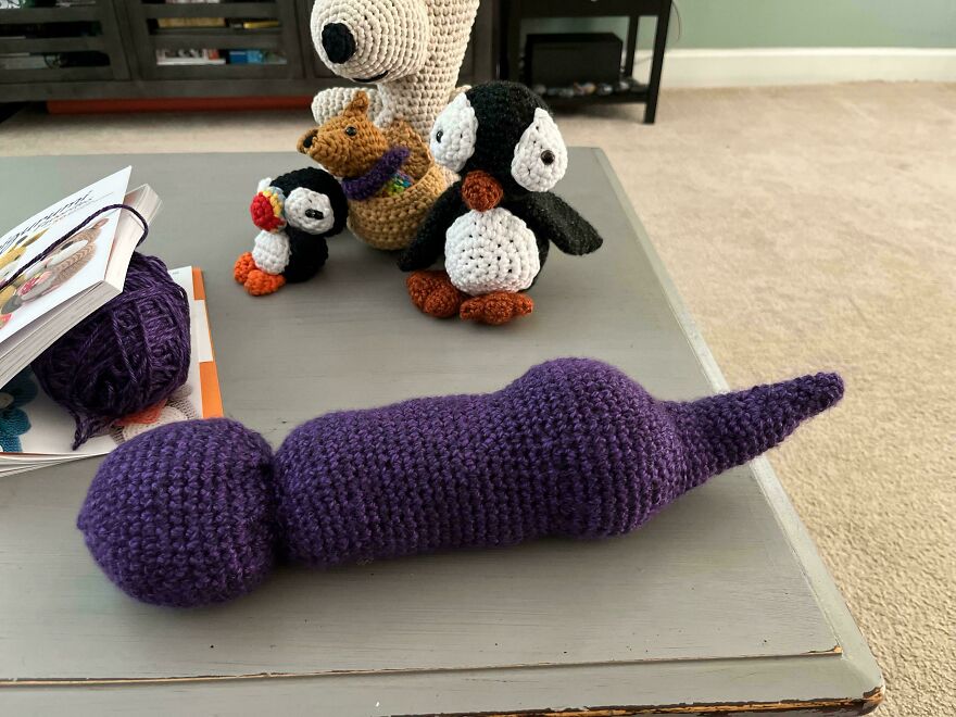 A purple crochet creation lies on a table next to a group of crochet penguins and other craft projects, awaiting completion.
