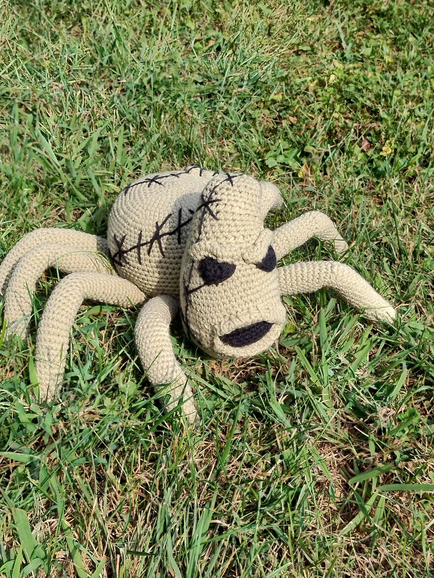 A large, tan crochet spider with stitched details and a menacing face rests in green grass, an example of crochet creations.