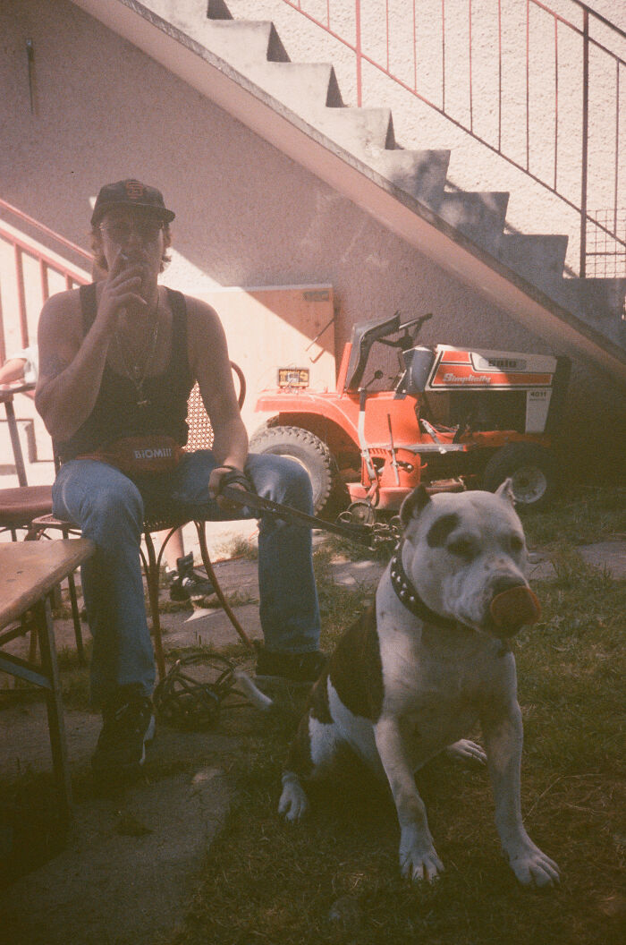 A man smoking with his dog sitting next to him, exploring abandoned places, with a tractor in the background.