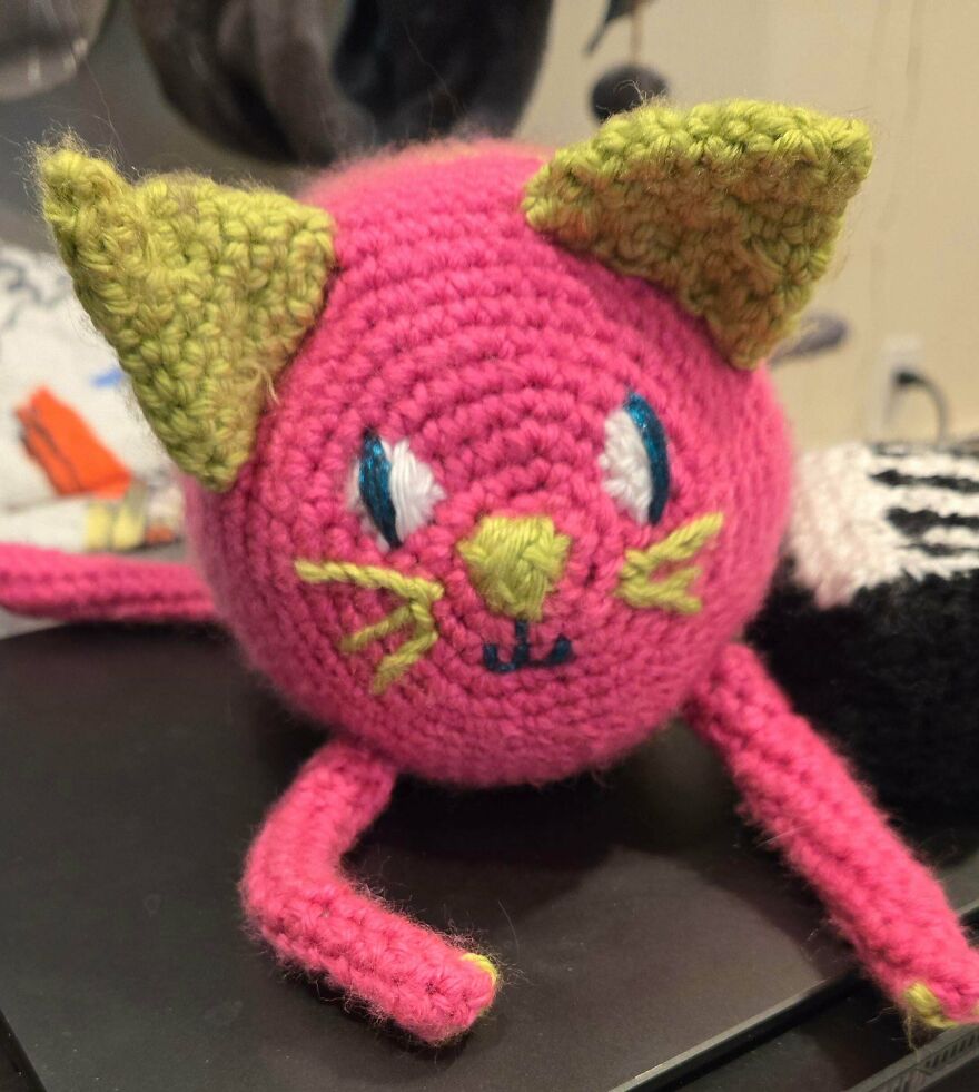 A pink crochet creation of a cat with green ears and blue eyes, a unique craft project.