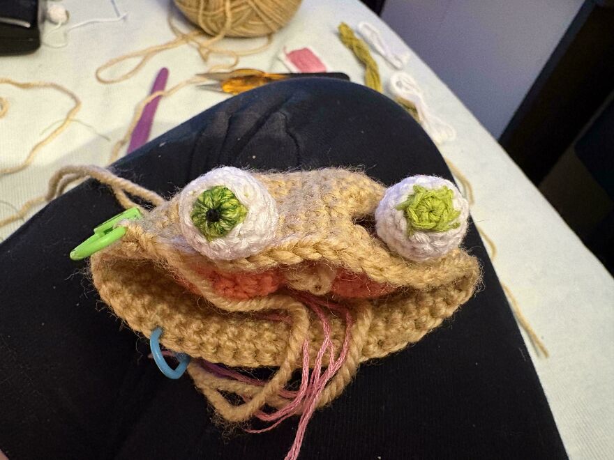 A close-up of a partially finished crochet creation with large green eyes and stringy pink and brown yarn.