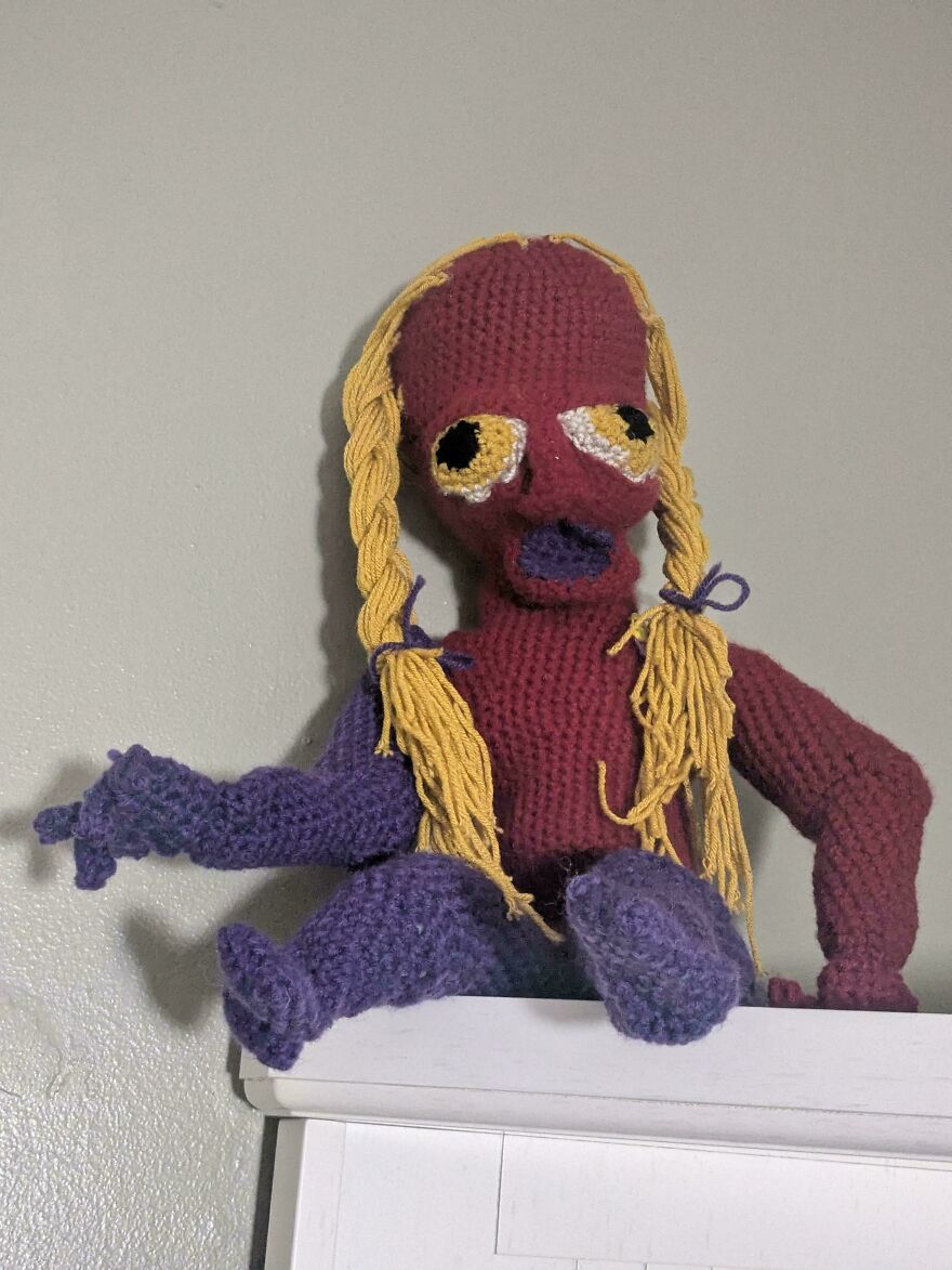 A creepy, maroon crochet creation doll with yellow pigtails and purple limbs, sitting on a white shelf.