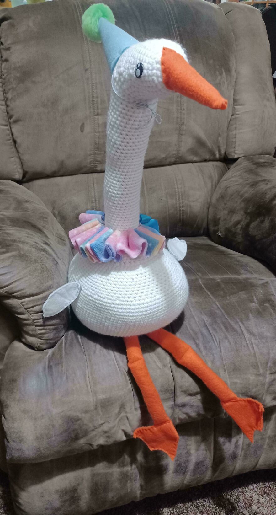 A large, white crochet creation goose wearing a party hat and a colorful ruffled collar, sitting on a brown chair.