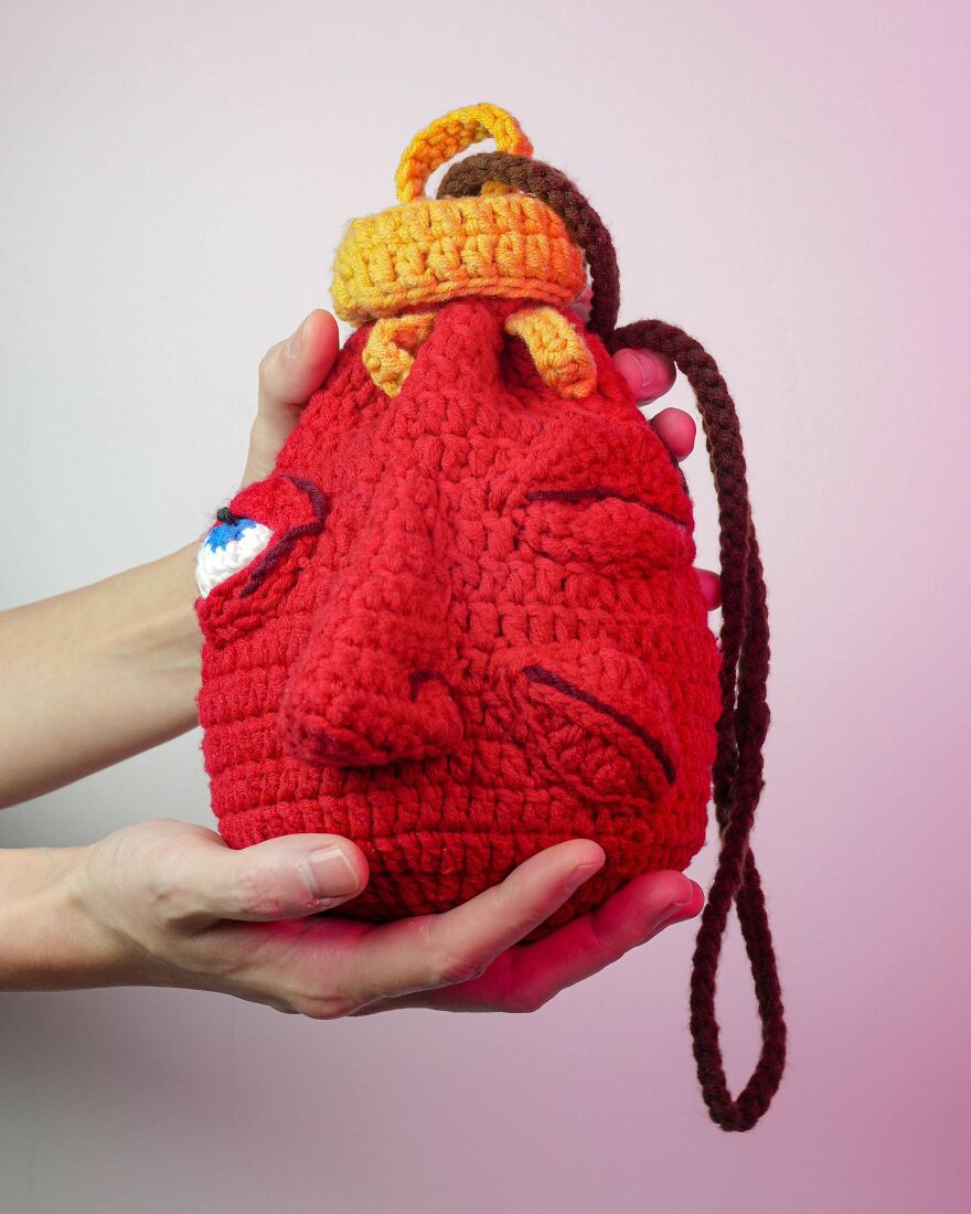 Hands holding a red crochet creation pouch resembling a face with an open mouth and visible white eye.