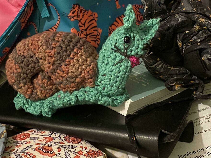 A crocheted snail with a green body and brown shell, featuring a cat's head, a funny craft project.