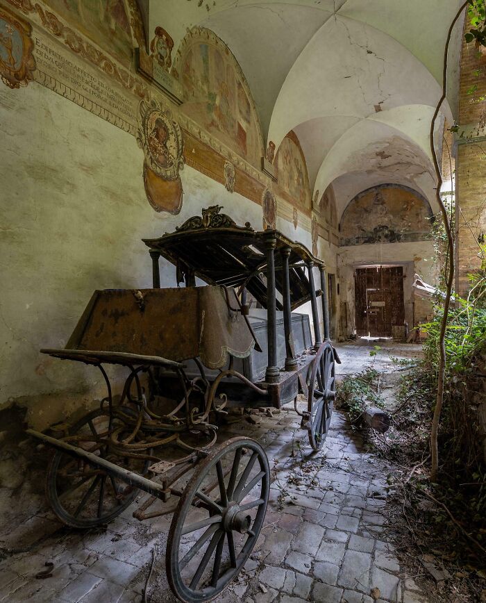 A vintage horse-drawn carriage sits in an overgrown, crumbling hall with painted walls in an abandoned place.