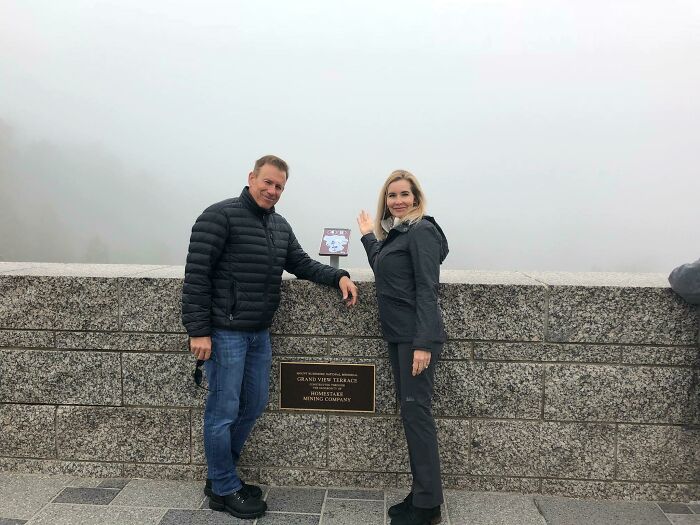 A man and a woman pointing at a small sign on a foggy overlook, illustrating a trip that didn't go as planned.