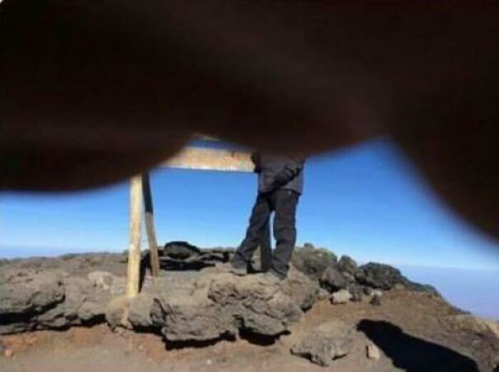 A poorly framed photo of a person standing on a rocky mountain peak against a clear blue sky, a trip mishap.