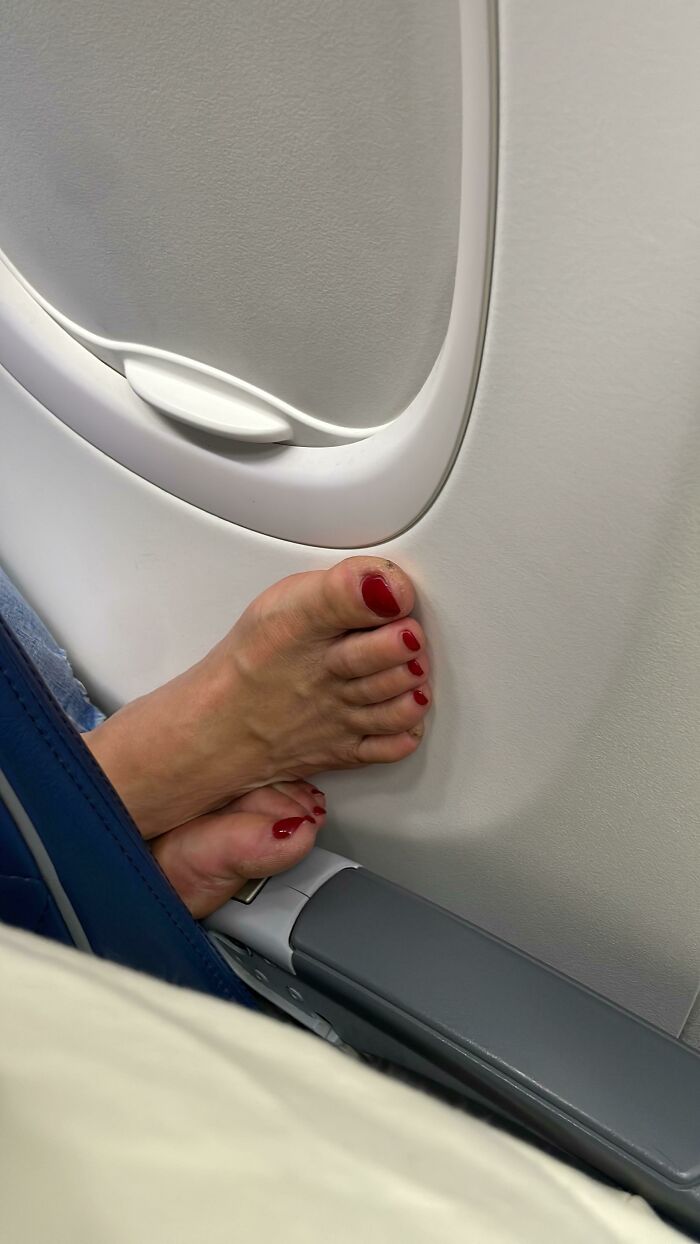 A person's bare feet with red nail polish resting against an airplane window, a travel trip annoyance.