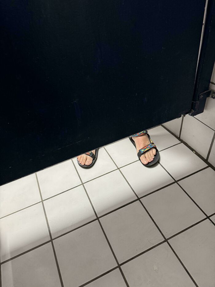 Feet in sandals visible under a restroom stall door, depicting entitled folks ignoring privacy.