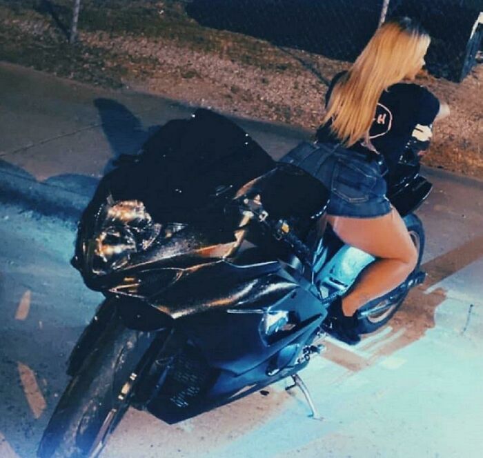 A woman with long blonde hair leaning over a black motorcycle. She is an entitled folk.