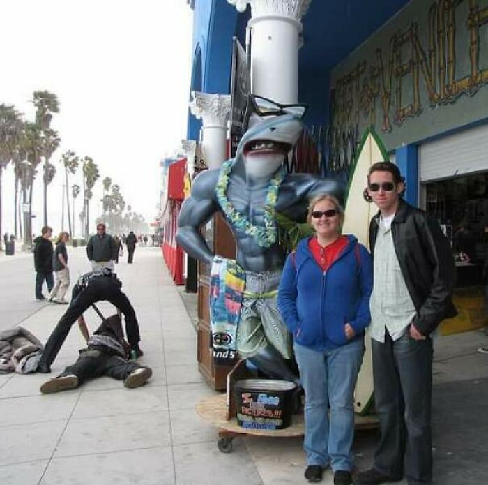A couple posing with a shark statue on a boardwalk, with a person bending over in the background, highlighting trips that didn't go as planned.
