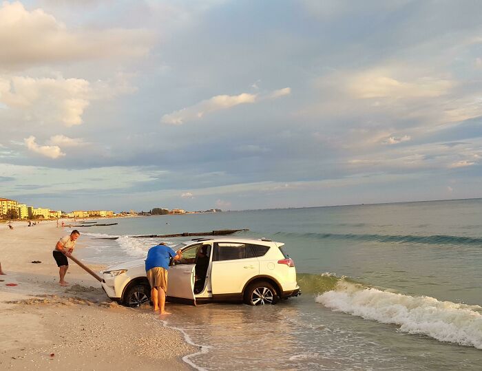 A white SUV stuck in the ocean with waves hitting its tires, showing people's trips gone wrong.