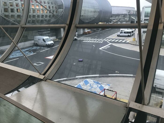 An airport tarmac seen through a geometric window, showing a single purple suitcase on the wet ground, symbolizing trips gone wrong.