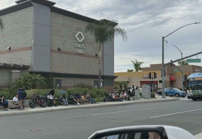Entitled folks waiting in a long line outside a Sam's Club, some seated on chairs, others standing.