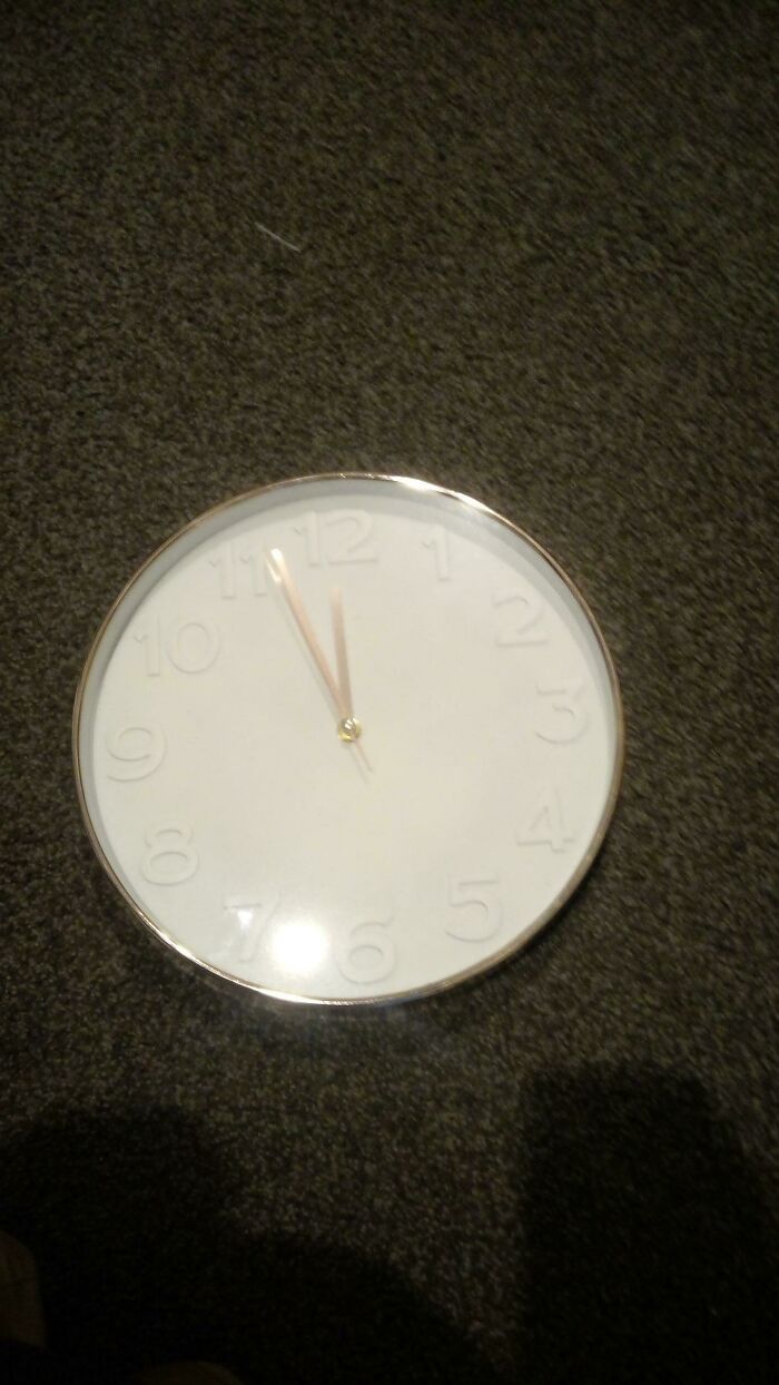 A white round clock with raised numbers and gold hands on a dark carpet, showing a design fail of legibility.