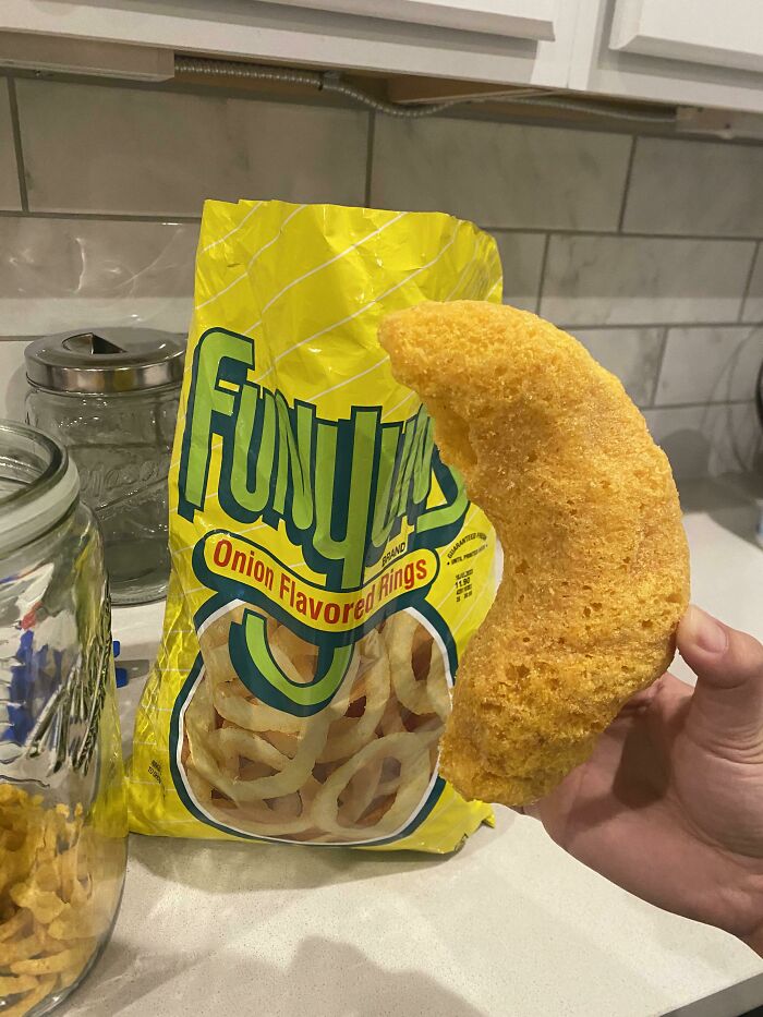 A hand holds a giant, oddly shaped Funyuns snack in front of its bag, a weird and unexpected moment.