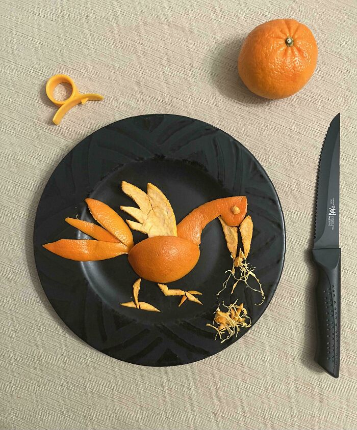 An artistic bird sculpture made from orange peels on a black plate, with a whole orange and knife nearby. A weird and interesting moment.