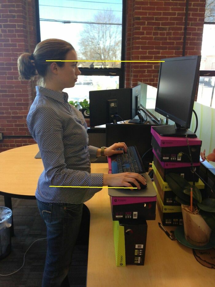 A woman at a DIY standing desk made of stacked shoeboxes, highlighting poor ergonomic setup.