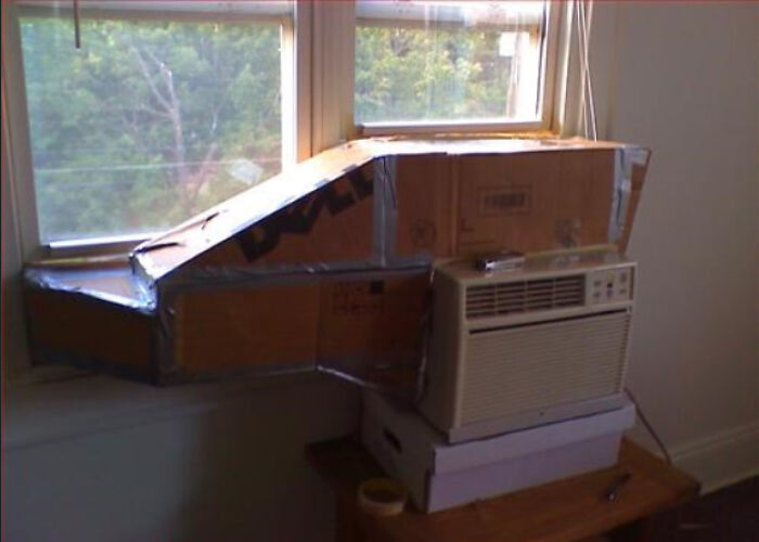 An air conditioner in a window with cardboard filling the gaps, a hilariously bad example of DIY.