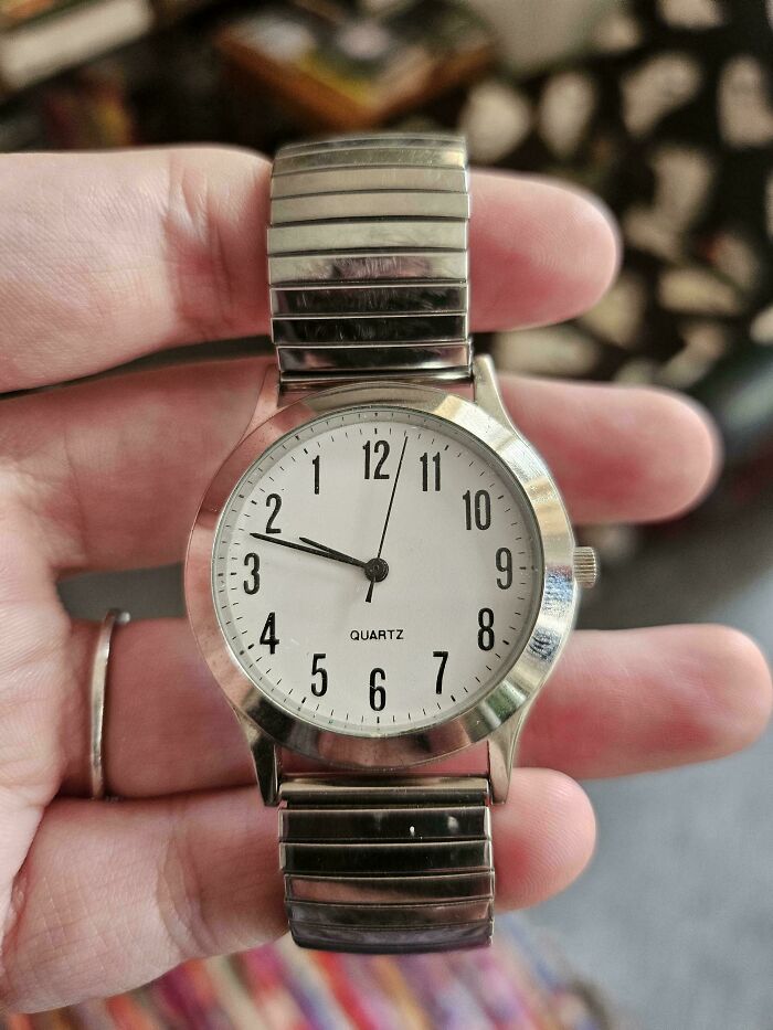A hand holding a simple silver watch with a white face and black numbers, revealing an unexpected moment.