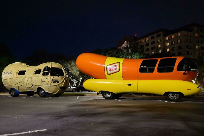 Two weird and unexpected vehicles, a Wienermobile and Planters Nutmobile, parked at night, making the day interesting.