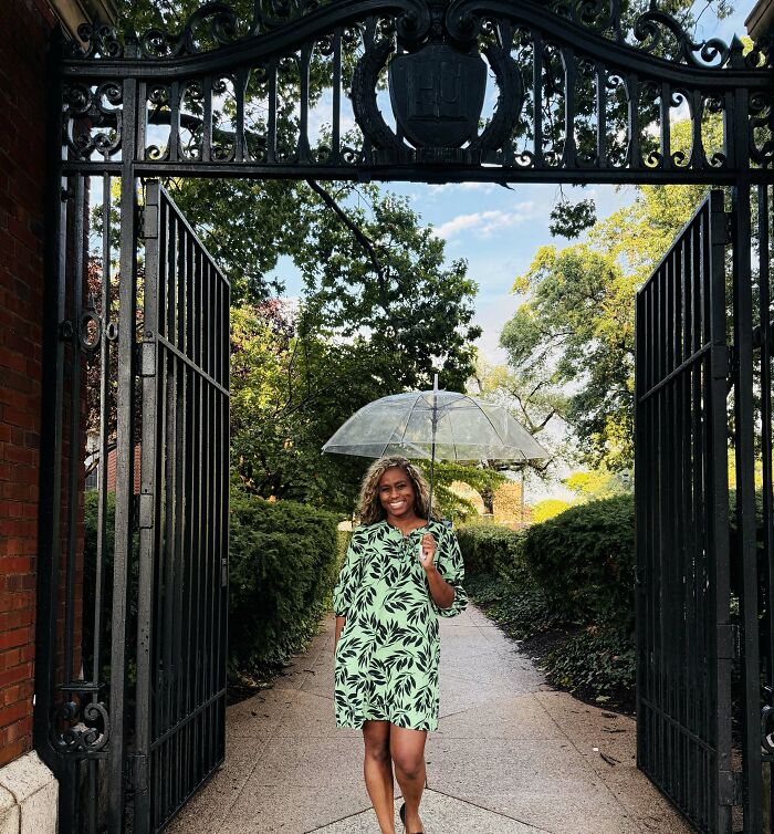 Smiling woman with curly hair walking through a large ornate gate with an umbrella, achieving incredible accomplishments