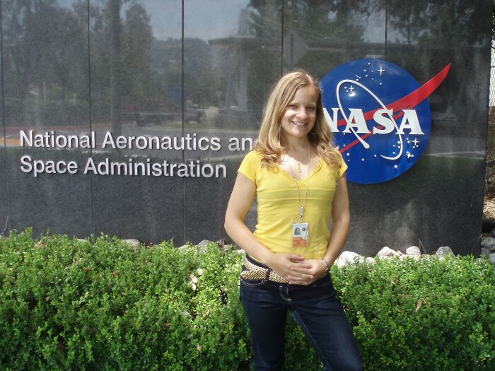 A smiling woman in a yellow shirt stands in front of the NASA sign, showcasing incredible accomplishments.