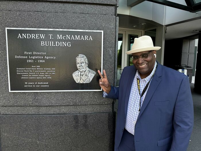 A man smiling and giving a peace sign next to the Andrew T. McNamara Building sign, celebrating his accomplishments.