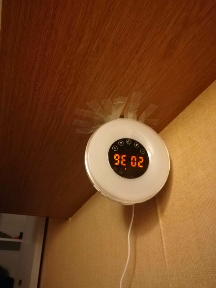 Hilariously bad DIY: a white circular alarm clock taped upside down to the underside of a wooden shelf, displaying 20:36.