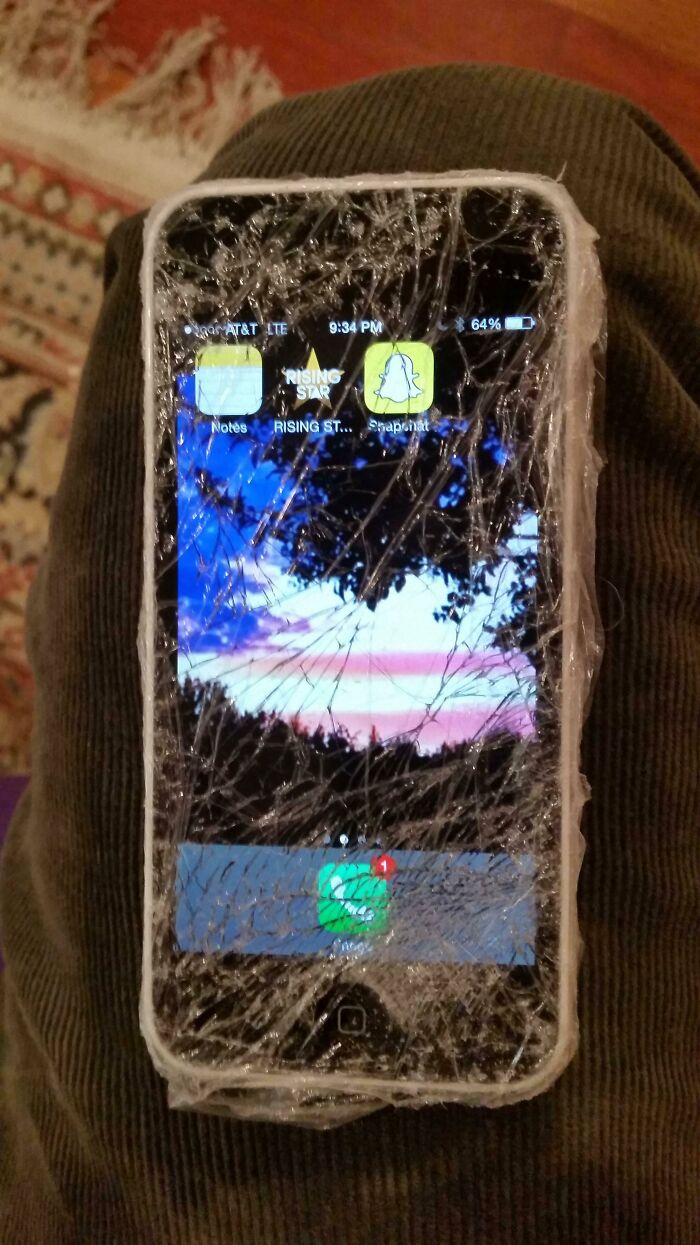 Hilariously bad DIY: a cracked iPhone screen, covered in clear tape, displaying app icons, held on a person's lap.